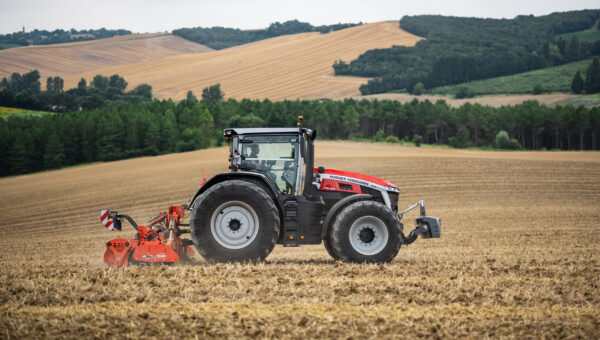 Side view of a Massey Ferguson 9S tractor cultivating a large field with rolling hills in the background